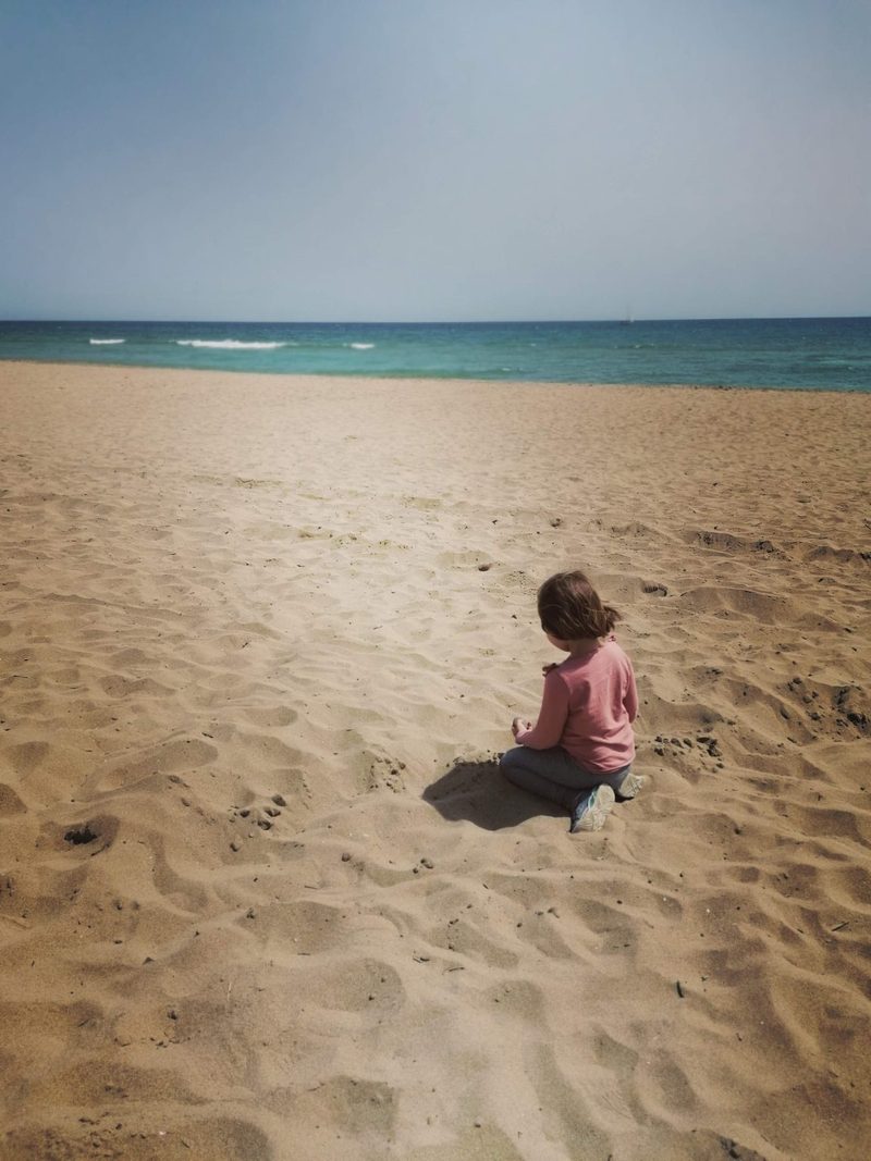 Ett barn sitter i sanden på en strand och tittar ut mot havet.